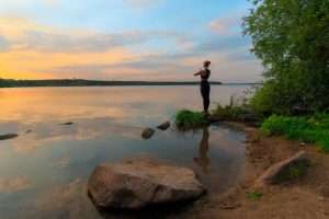 A woman standing in Namaste pose on a lakeshore at sunset, symbolizing embodied gratitude, inner peace, and the quiet rebellion of healing after trauma.
